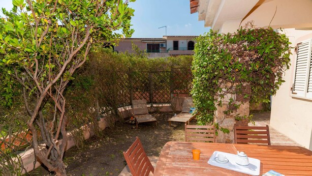 Un patio con mesa y sillas de madera, un pequeño jardín con un árbol y un edificio al fondo.