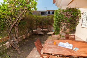 A patio with a wooden table and chairs, a small garden area with a tree, and a building in the background. - Fantastic Sardinia Casa Piras pir01 (Budoni)