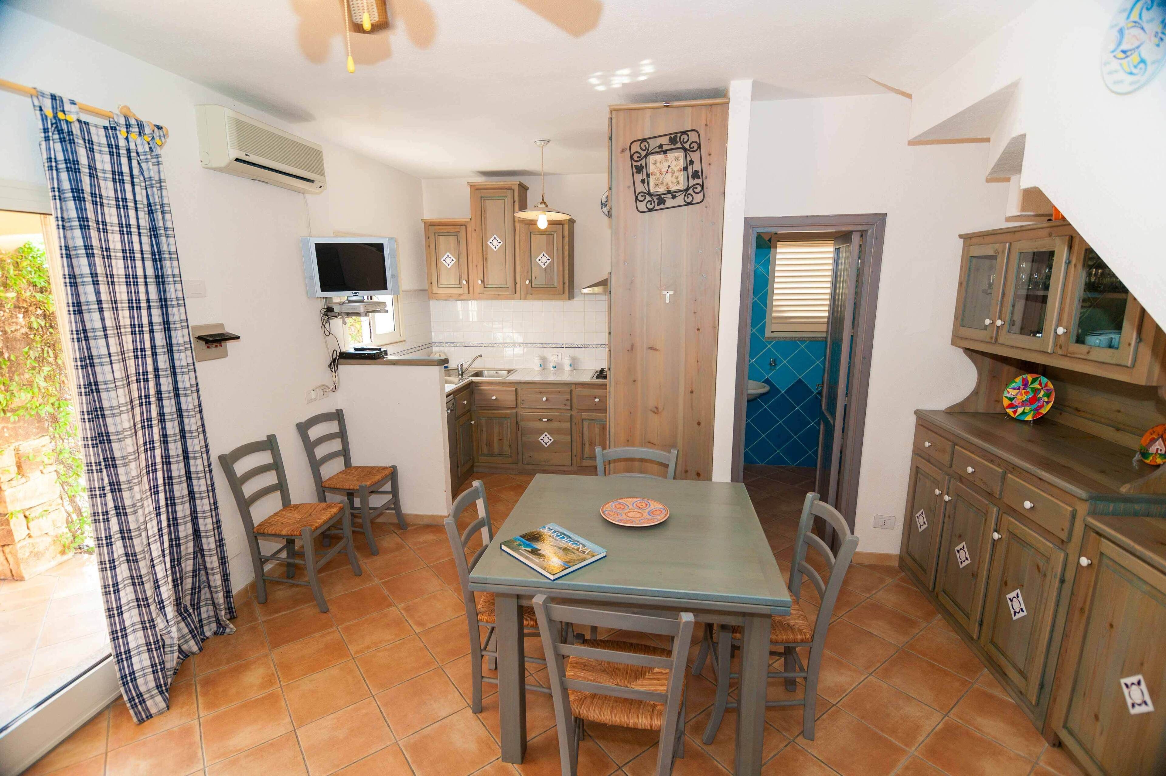A kitchen with wooden cabinets, a dining table with chairs, a TV, and a blue door.