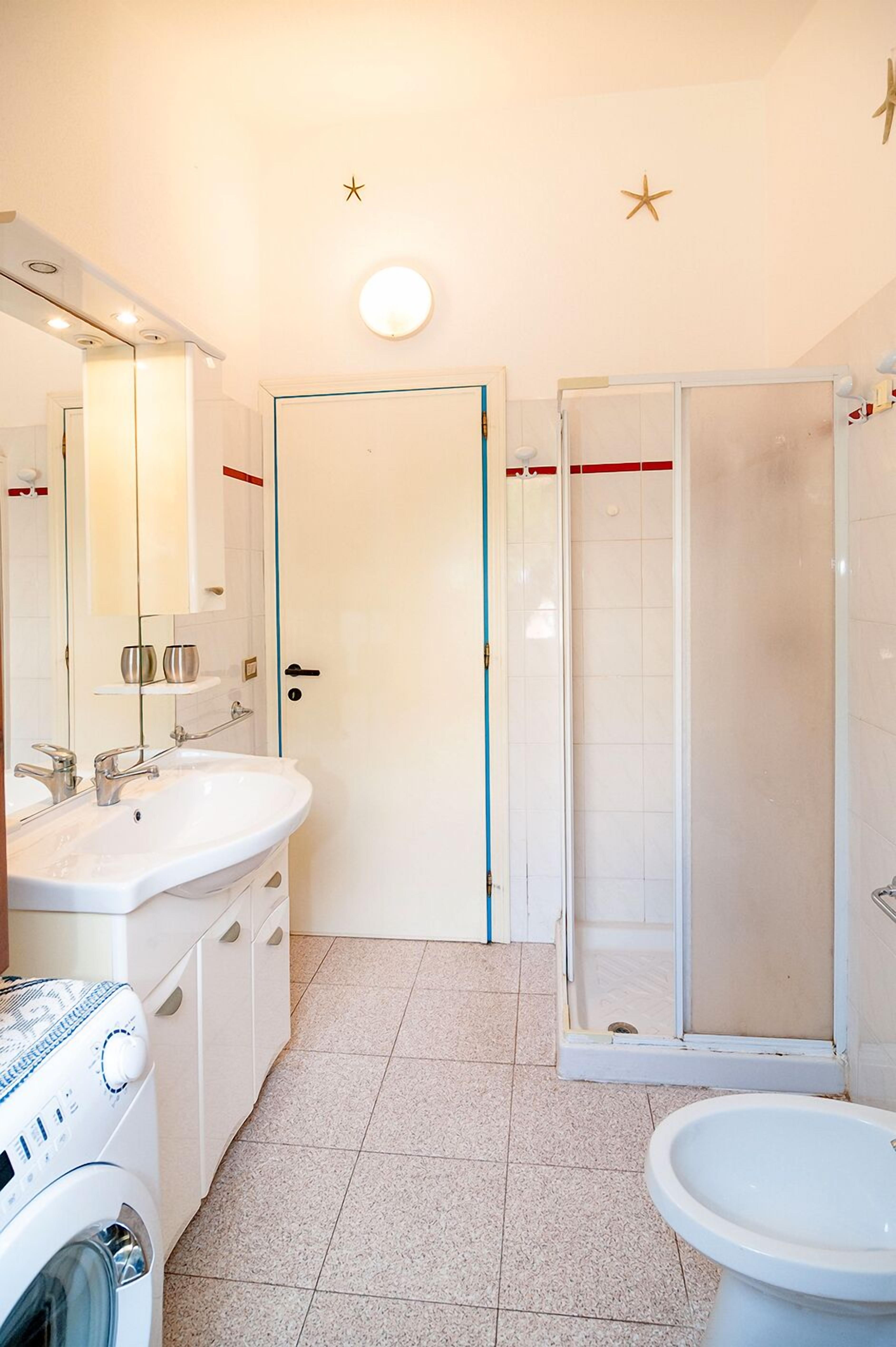 Et badeværelse med to håndvaske, toilet, vaskemaskine og en brusekabine i glas.