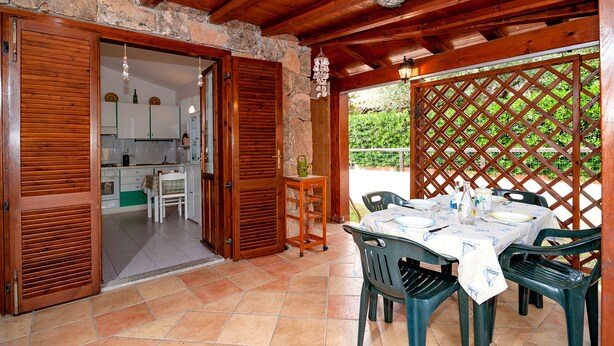 A covered patio area with a table set for dining, wooden doors, and a kitchen visible through an open door. - Fantastic Sardinia Casa Podas pod01 (Budoni)