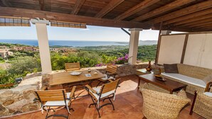 An outdoor patio with a wooden table and chairs, a wicker sofa, and a stone wall. - Fantastic Sardinia Casa Castas cast01 (Budoni)