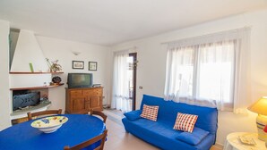 A living room with a blue sofa, a dining table with chairs, a television on a wooden cabinet, and a window with sheer curtains. - Fantastic Sardinia Casa Castas cast01 (Budoni)