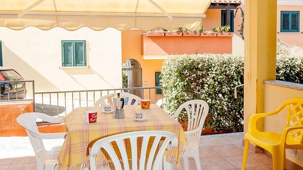 Un espace extérieur abrité, aménagé pour prendre les repas, avec une table et des chaises, un parasol jaune et une chaise jaune.