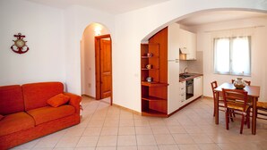 Un espace de vie compact comprenant un canapé, une table à manger avec des chaises, un coin cuisine et une porte en bois donnant sur une autre pièce.