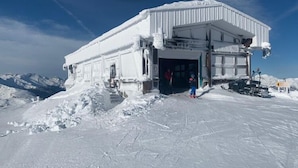 Ein schneebedecktes Gebäude mit einem großen Eingang, vermutlich eine Berghütte oder Station, umgeben von Schnee und Bergen im Hintergrund.