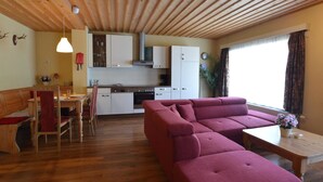 Ein gemütliches Wohnzimmer mit Holzbalkendecke, einem roten Sofa, einer Essecke mit Holztisch und -stühlen sowie einer Küche mit weißen Schränken und einem Herd.