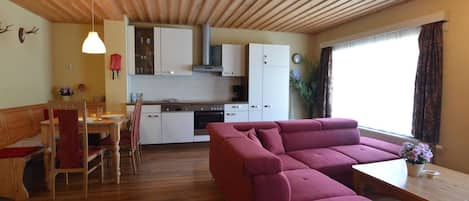 Una sala de estar acogedora con techo de madera, un sofá rojo, un comedor con mesa y sillas de madera, y una cocina con armarios blancos y una estufa.