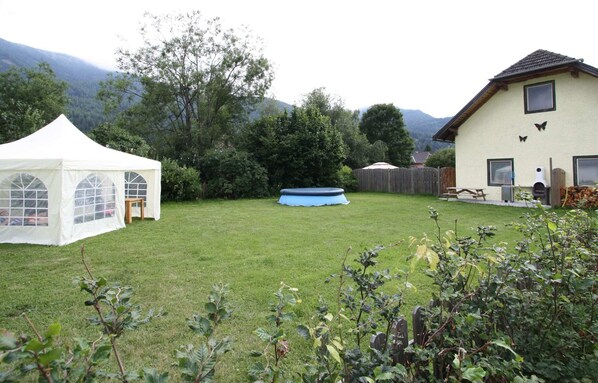 Un prato verde con una tenda bianca, un tappablocchi blu e una casa con le montagne sullo sfondo.