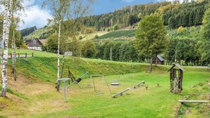 Une aire de jeux avec des balançoires et des structures d’escalade, entourée d’une pelouse avec des arbres et une cabane en bois en arrière-plan.