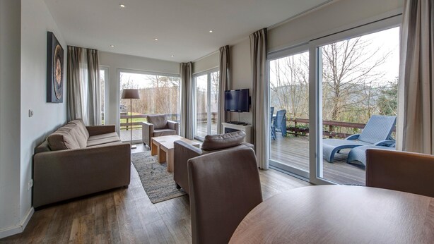 A modern living room with a sofa, chairs, a coffee table, and a TV. Large windows offer a view of the outdoors. - Detached Bungalow with Washing Machine near Lake (Dahlem)