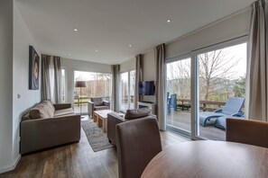 A modern living room with a sofa, chairs, a coffee table, and a TV. Large windows offer a view of the outdoors. - Detached Bungalow with Washing Machine near Lake (Dahlem)
