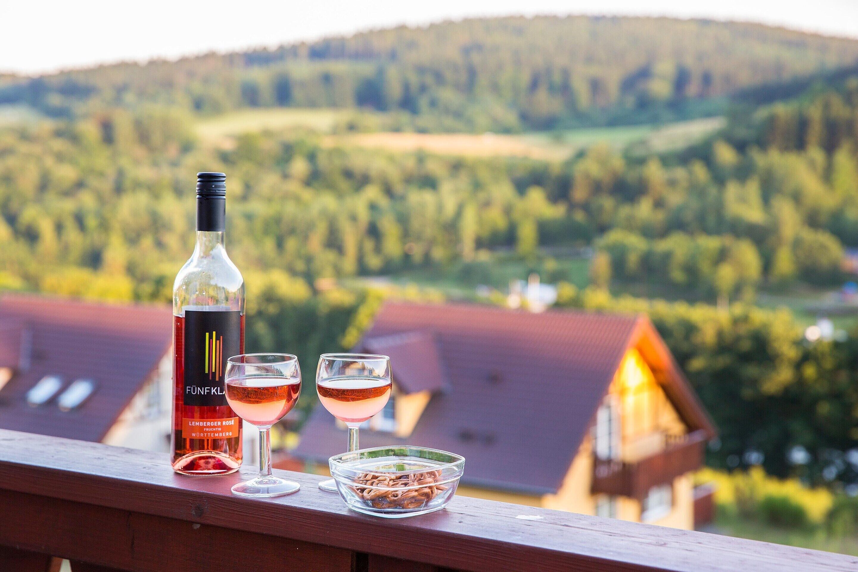 A bottle of wine and two glasses on a wooden railing with a view of a house and hills in the background.