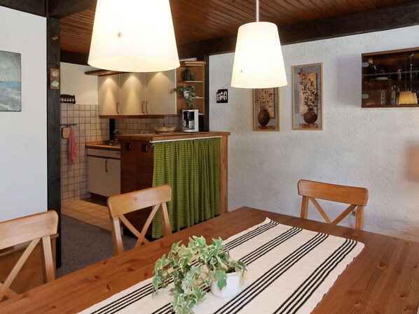 A dining area with a wooden table, chairs, and a green table runner. There is a kitchen in the background with a tiled backsplash and wooden cabinets. - Wooden Chalet with Terrace near Swimming Lake (Waldkirchen)