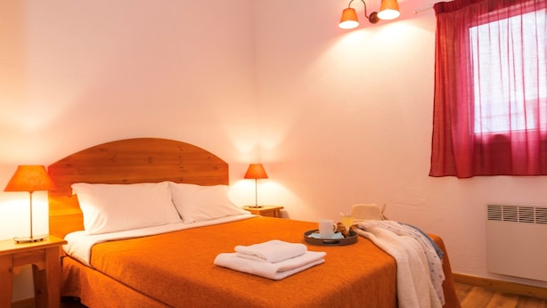 Uma cama bem arrumada com cabeceira de madeira, duas luminárias de apoio, um tabuleiro com chávena e pastelaria, e uma janela com cortina vermelha.