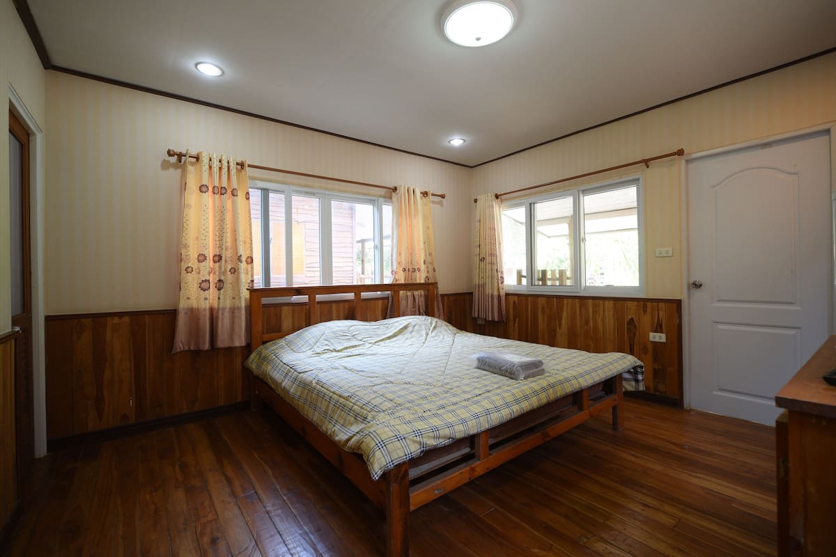 Ein Schlafzimmer mit einem hölzernen Bett, zwei mit Vorhängen versehenen Fenstern, einer Tür und einer Deckenleuchte.