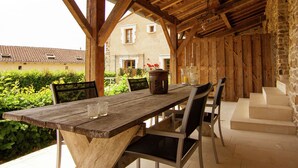 A wooden table with chairs under a wooden roof, set against a stone wall with a house in the background. - Gorgeous Holiday Home in Roussines With Terrace (Roussines)