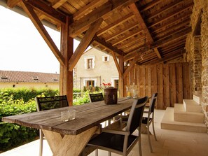 A wooden table with chairs under a wooden roof, set against a stone wall with a house in the background. - Gorgeous Holiday Home in Roussines With Terrace (Roussines)