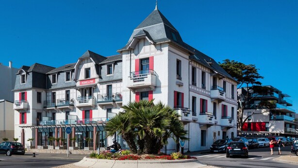Un hotel de varios pisos con balcones, un patio central con palmeras y un cielo azul despejado.