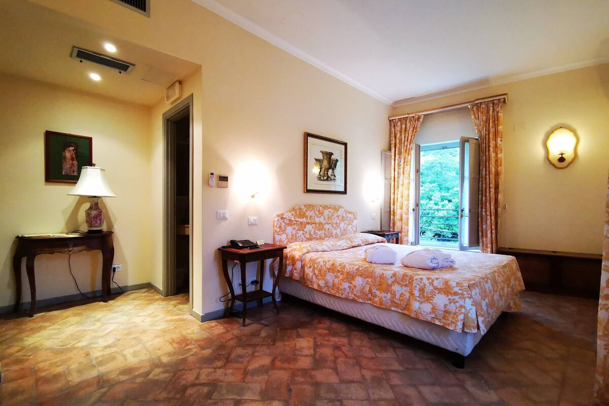 Ein Schlafzimmer mit einem Bett, einem Nachttisch, einer Lampe, einem Bild an der Wand und einem Fenster mit Vorhängen.
