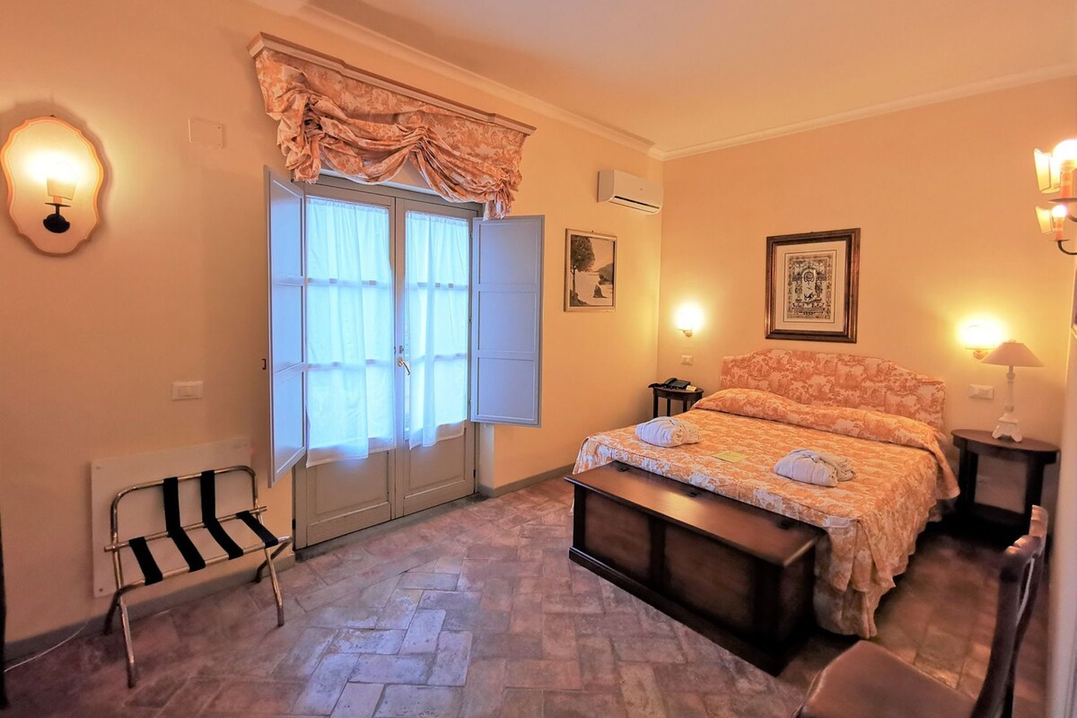 Une chambre à coucher comprenant un lit, des tables de chevet, un banc, une chaise, une fenêtre avec des rideaux et des lampes fixées au mur.