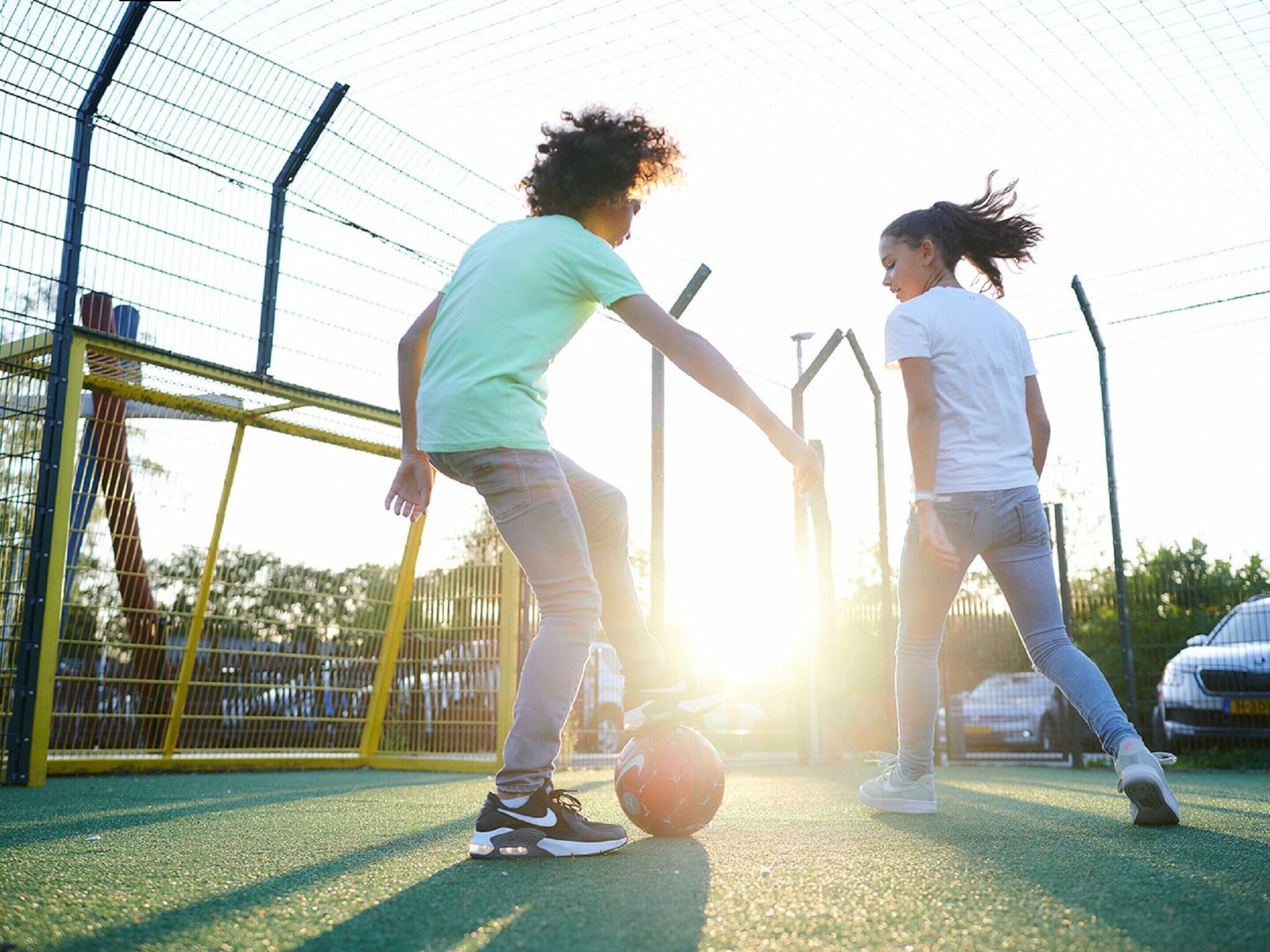 Two people playing soccer on a green field with a goalpost and a fence in the background.