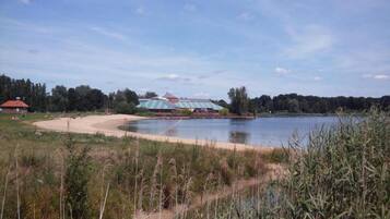Zona junto a un lago con un edificio de techo a rayas, rodeado de árboles y una playa de arena.