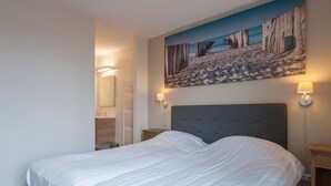 Une chambre d’hôtel avec un grand lit, une salle de bain équipée d’un porte-serviettes et un tableau représentant une scène de plage accroché au mur.