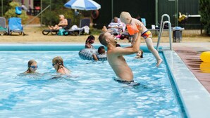 Piscina com pessoas na água, posto de salva-vidas e área cercada com cadeiras e guarda-sóis.