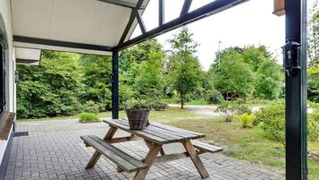 An outdoor patio with a wooden picnic table and benches, surrounded by a brick floor and greenery.