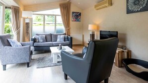 Ein modernes Wohnzimmer mit einem grauen Sofa, einem schwarzen Sessel und einem weißen Couchtisch. Ein Fernseher steht auf einem Holzgestell, und an der Wand hängt ein Gemälde.