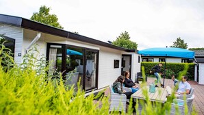 Ein moderner Außenbereich mit einer Terrasse, Korbmöbeln, einem Glastür-Eingang und einem blauen Sonnenschirm.