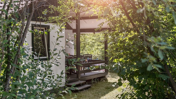 Une terrasse en bois attenante à un bâtiment blanc doté d’un toit en tuiles, le tout entouré de verdure.