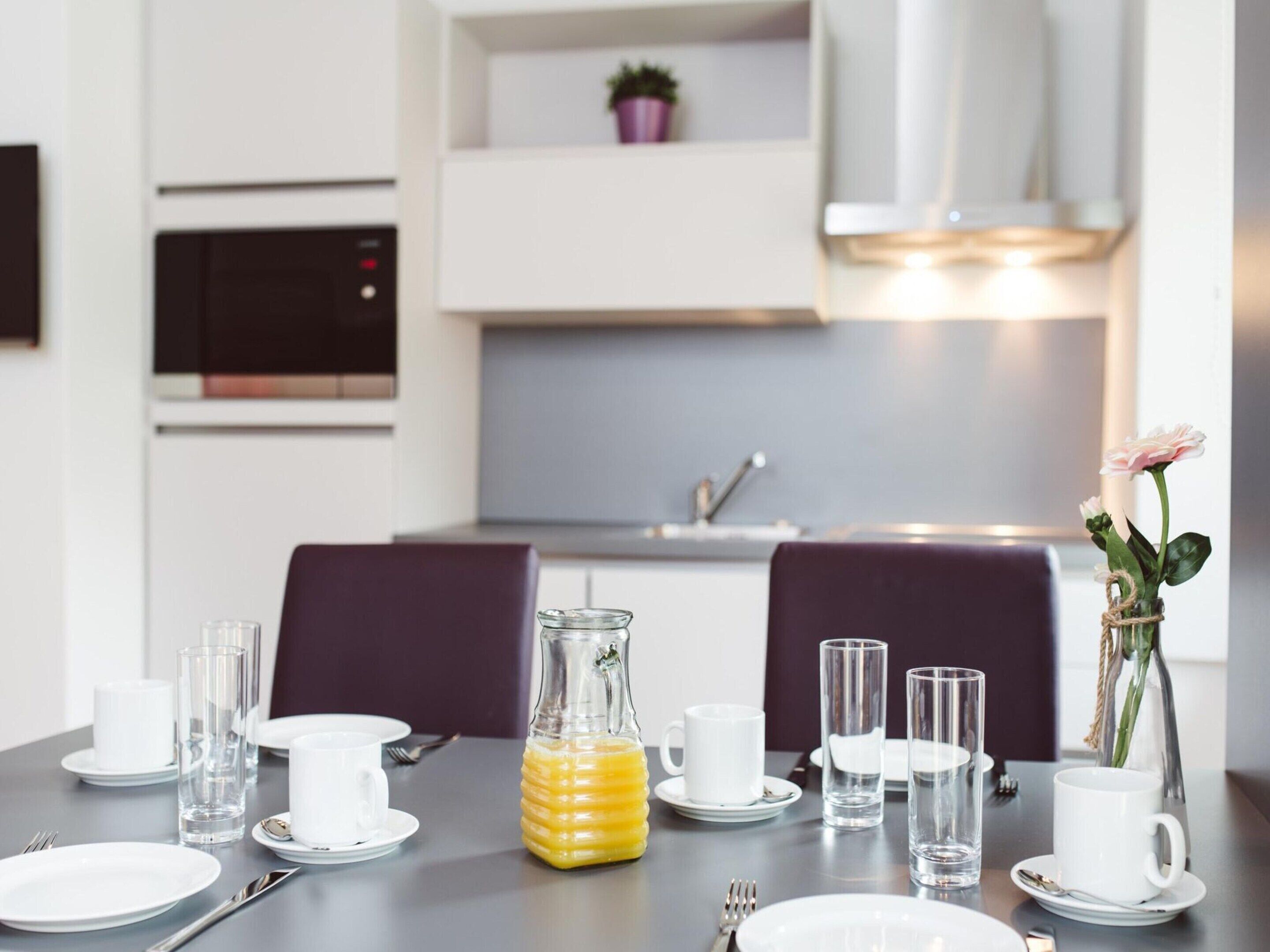 A modern dining area with a table set for two, featuring white plates, clear glasses, and a bottle of orange juice.