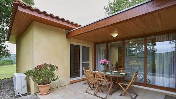 Une terrasse avec une table et des chaises, un pot de fleurs et une porte-fenêtre coulissante donnant sur un espace intérieur.