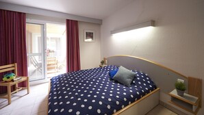 Une chambre à coucher comprenant un lit, une chaise, une petite table, une plante en pot et donnant sur un balcon extérieur.