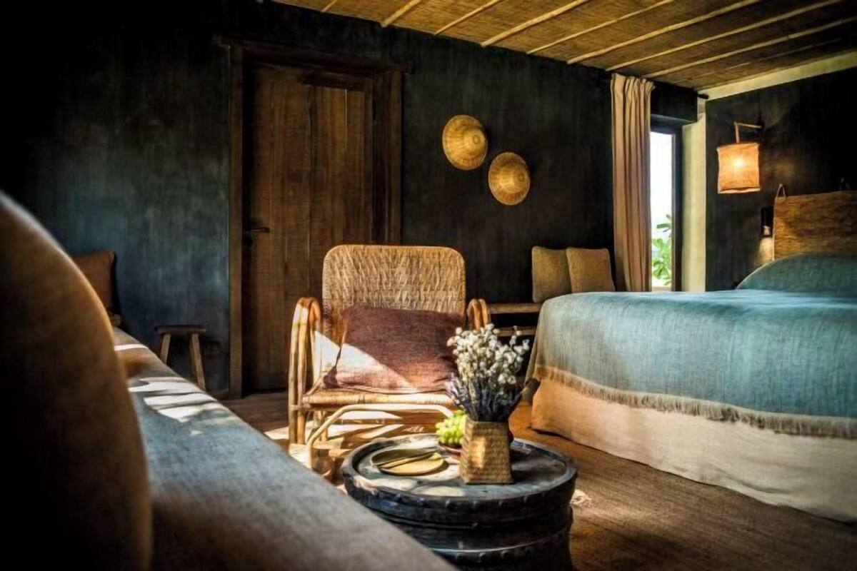 Ein gemütliches Zimmer mit einem Bett, einem Korbstuhl, einem runden Tisch mit einer Vase und einer Holztür.