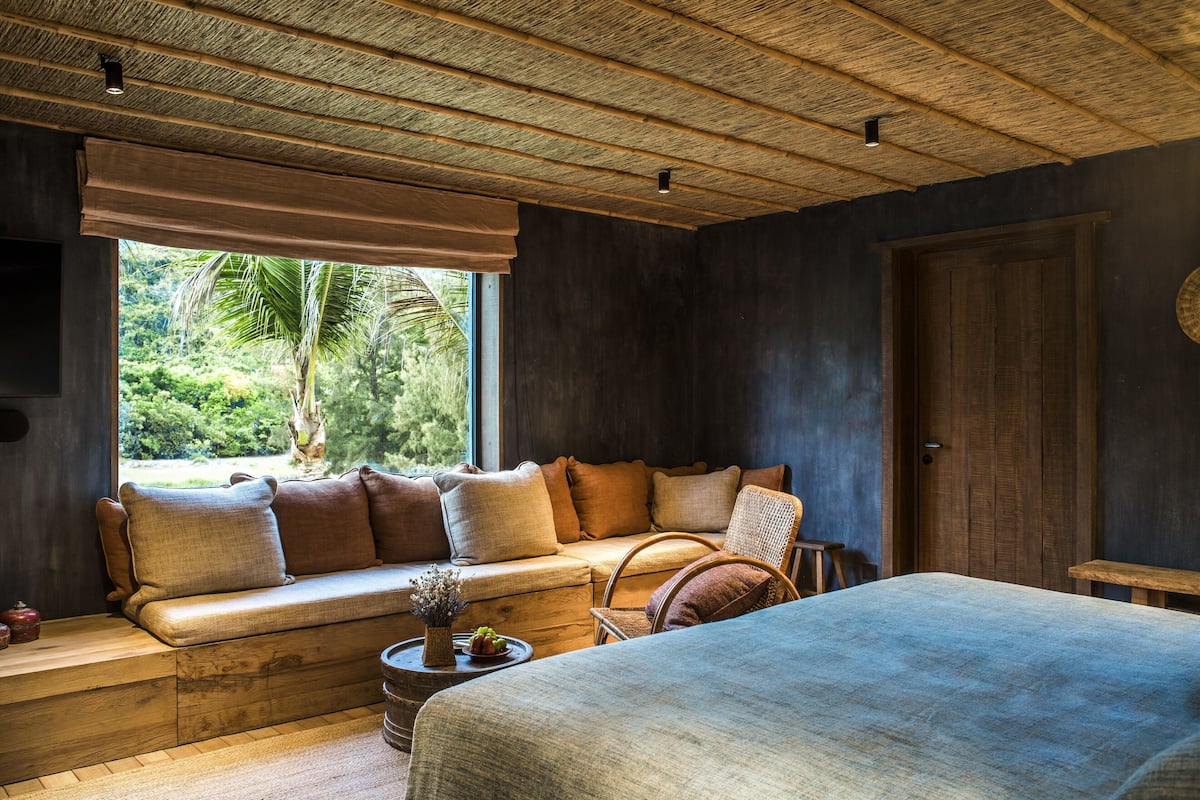 Ein Zimmer mit einem großen Bett, einer Couch mit Kissen, einer hölzernen Bank und einem Fenster mit Blick ins Grüne.