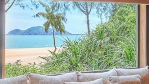 A room with a large window overlooking a beach, a sofa with cushions, and a small wooden table with a bowl of fruit. - Zannier Bãi San Hô (Xuan Canh)