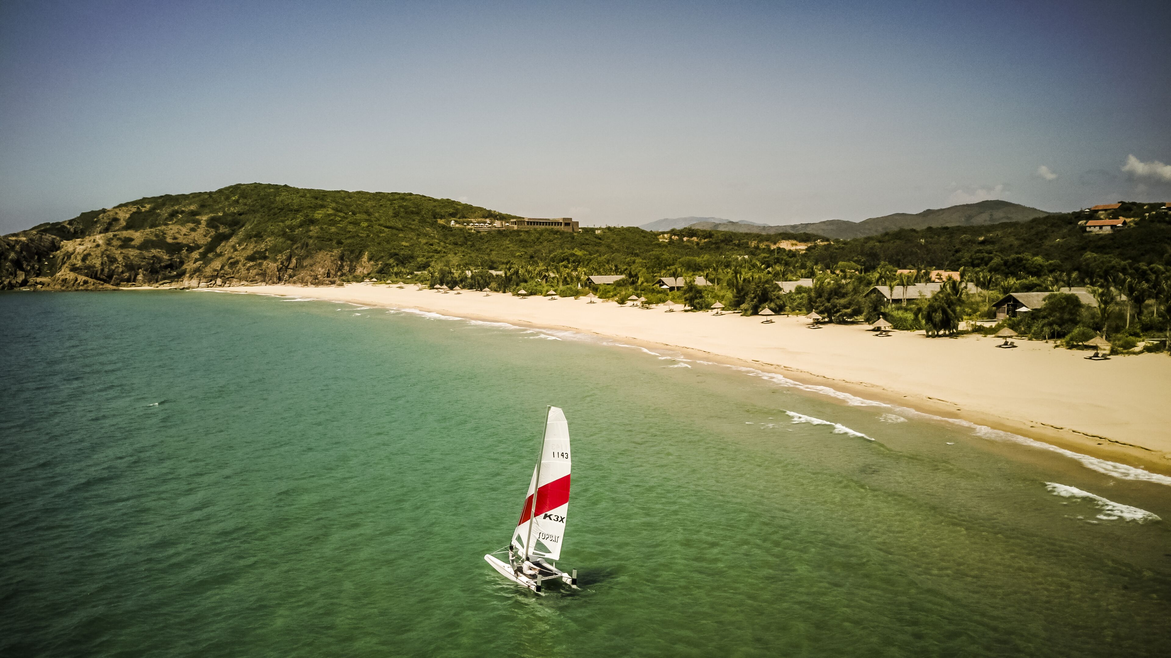 A sailboat on a clear turquoise sea with a sandy beach and greenery in the background.
