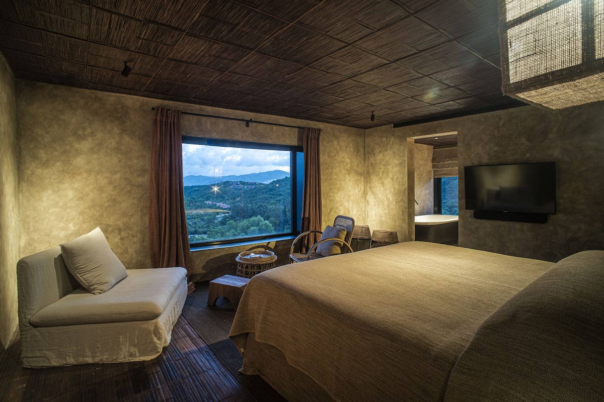 Ein Hotelzimmer mit einem großen Bett, einer Couch, einem Fernseher und Blick auf eine Berglandschaft durch ein Fenster.