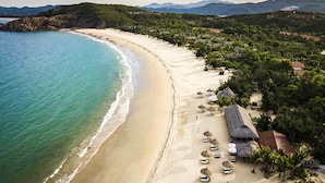 A beach with a long stretch of sand, a few umbrellas, and a few buildings in the background. - Zannier Bãi San Hô (Xuan Canh)