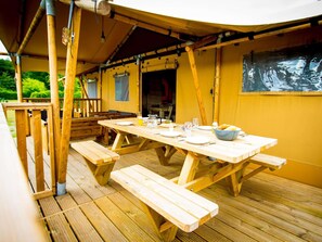 Une terrasse en bois avec des tables de pique-nique et des bancs, un espace couvert aux murs jaunes, et une peinture murale.