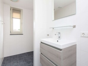 Ein modernes Badezimmer mit weißen Fliesen, einem dunklen Duschbereich, einem weißen Waschbecken mit Spiegel darüber und einem Fenster mit cremefarbener Jalousie.