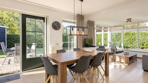 A modern dining area with a wooden table and chairs, a clock on the wall, and a view of the garden through large windows. - Holiday Home in Park in Twente (Hoge Hexel)