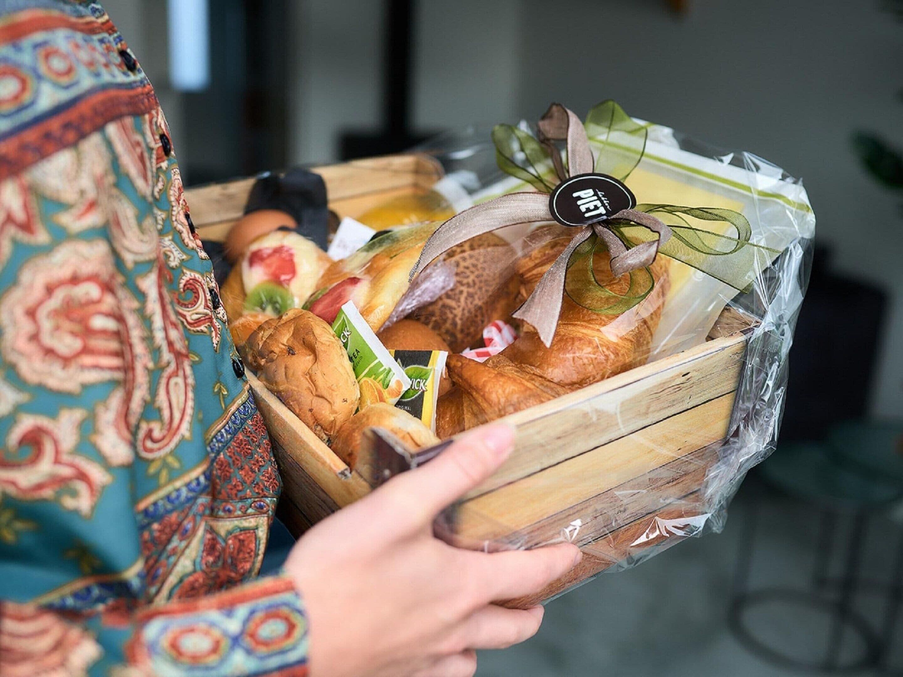A person holding a wooden basket filled with various breads and pastries, wrapped in clear plastic with a ribbon and a tag.