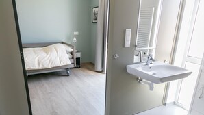 A bedroom with a bed, bedside table, lamp, and a bathroom with a sink and mirror. - Bungalow in Netherlands With Accessible Design ('s-Gravenzande)