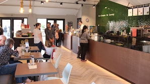 A modern café interior with wooden tables, white chairs, and a green wall with menu options. - Cozy Lodge in the Achterhoek (Winterswijk - Brinkheurne)