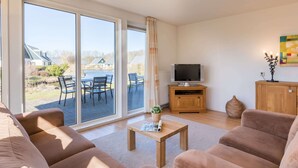 Un salon comprenant un ensemble de canapés, une table basse en bois, un téléviseur posé sur un meuble, et une vue sur une terrasse avec des chaises et une table.