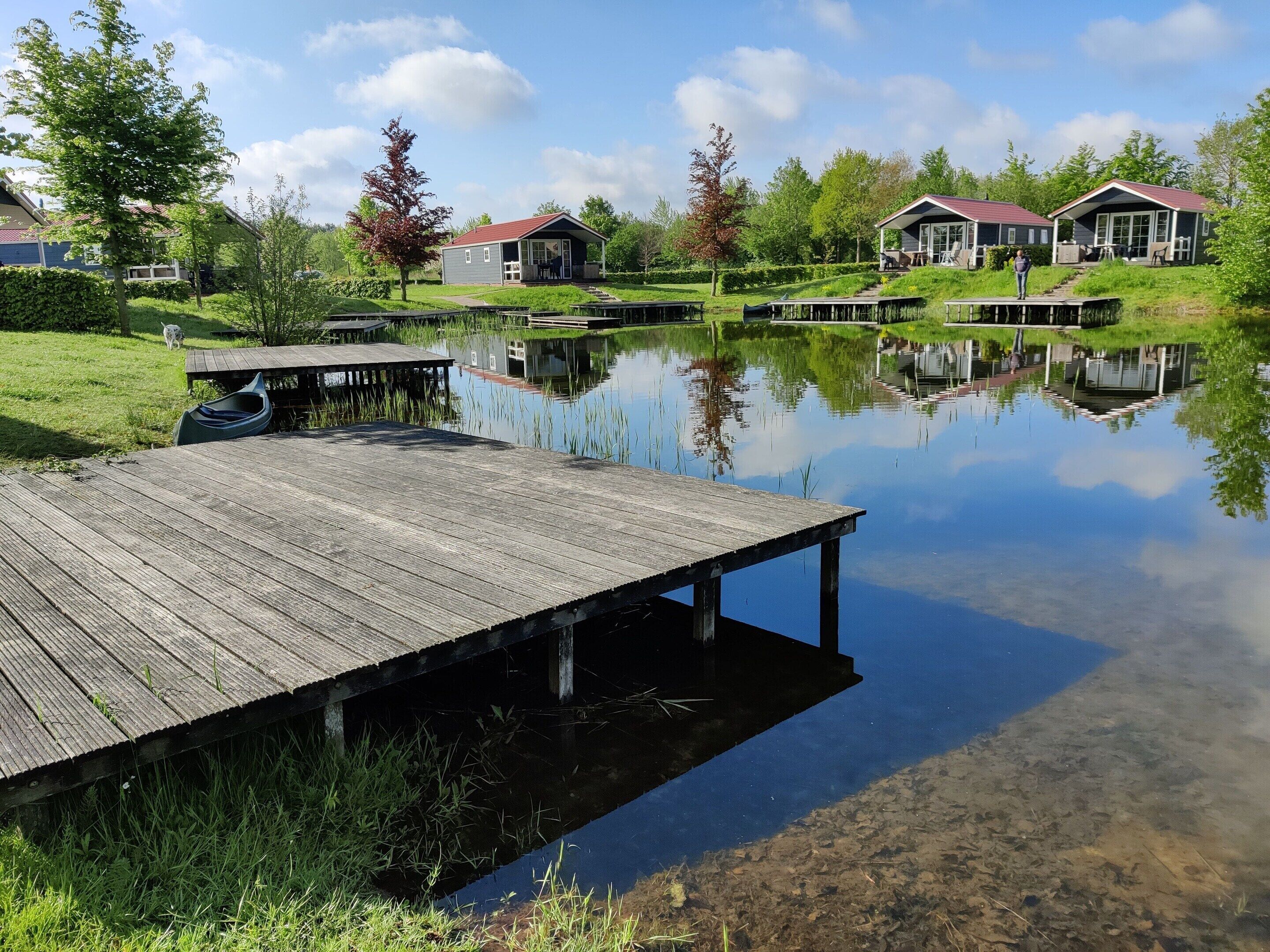 A wooden dock extending into a calm lake, surrounded by lush greenery and small houses with red roofs.